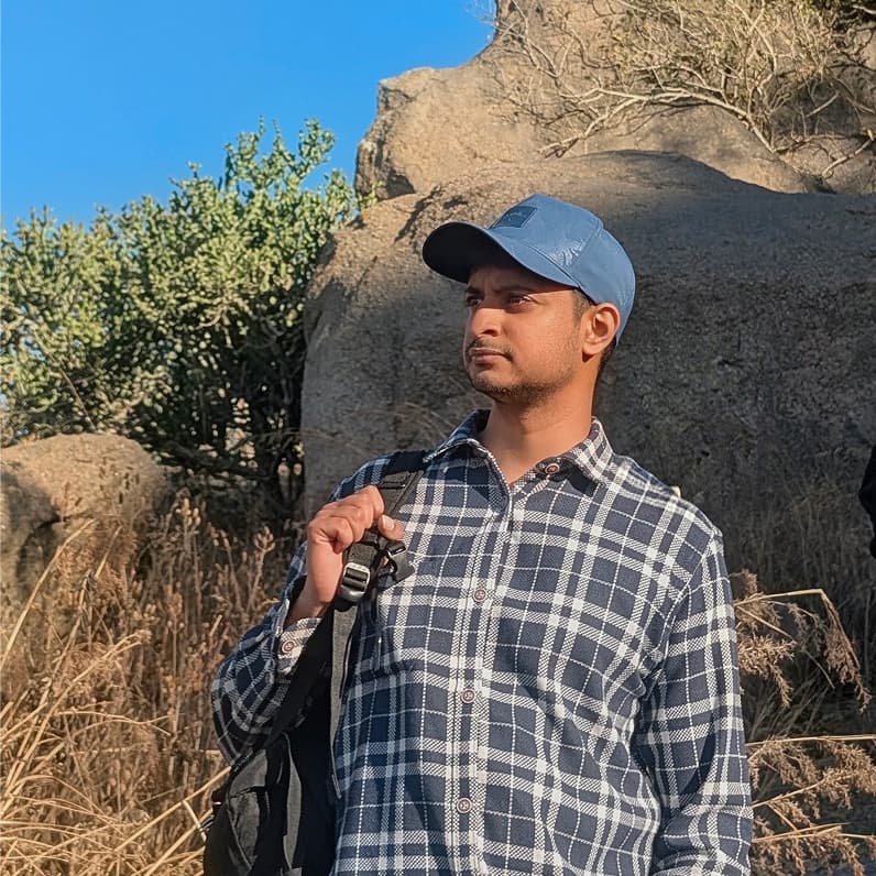 Shiv Dinesh in a blue cap and checked shirt, standing among sunlit boulders and dry grass